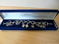 Full view of the sterling silver link charm bracelet held in a blue velvet box with white interior lid that reads 'DeForest'. All charms are attached to the bracelet.