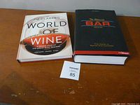 Two hardcover books on a wooden surface: The Ultimate Bar Book with black cover and World of Wine by Oz Clarke with a wine glass image on the cover.