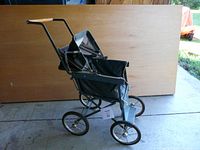 Side view of vintage metal doll carriage showing adjustable hood, black and blue fabric seating, metal wheels with rubber, and handlebar with wooden grips.