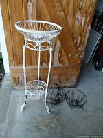 Full view of white metal plant stand with two tiers and five black wire hanging baskets placed beside it on the floor.