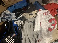 Photo showing a variety of men's clothing items including a white sports jersey with red lettering (number 3), grey Polo by Ralph Lauren quarter-zip sweatshirt, black hoodie with NBA patch, and assorted other tops.