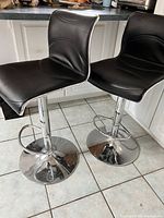 Two black vinyl and chrome bar stools positioned side by side on a tiled kitchen floor.