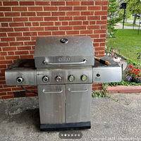 Full front view of stainless steel BBQ grill with four control knobs, two-door storage cabinet, side shelves, and built-in thermometer on lid.