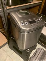 Bread maker on shelf with stainless steel body, control panel, and viewing window on top.