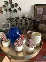 Metal candle holder with 5 glass votive holders, ceramic pots with artificial succulents, blue glass orb, textured ceramic candle holder, and other decor items on a hammered metal round tray with floral and framed wall decorations in background.