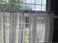 Four sheer white lace style curtain panels hung on a dark wooden rod in front of a large window, showing their delicate branch pattern.