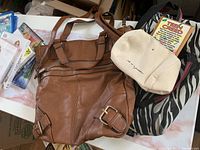 Large brown leather bag with buckle detail and handles placed beside small beige purse and teen creed book along with zebra patterned fabric.