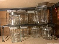 Front angled view of 10 clear glass jars resting on two metal shelves, showing variety of sizes and shapes with plastic and metal lids.