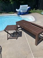Outdoor bench and matching small table shown on poolside concrete surface. Bench is rectangular with multiple wood composite slats, one slat missing. Small table is square with foldable legs. Both brown in color.