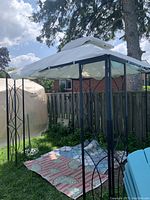 Full view of outdoor gazebo with black metal frame and cream/beige fabric canopy. Patterned outdoor rug under the gazebo.