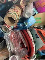 Box with several rolls of colorful gift wrapping paper, some clear disposable plastic cups, red and white plastic cutlery, and other party supply items.