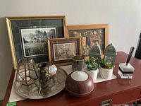 Photo of small framed paintings on row behind an assortment of decorative items including metal lanterns, faux succulents, metallic vase, geometric candle holders, round wooden diffuser, and reed diffuser.