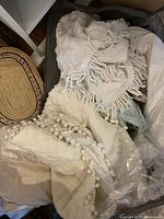 Overview of vintage linens including textured fabrics with pom-pom and tassel details, folded and arranged in a container.