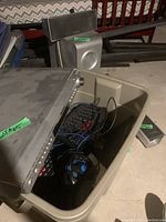 Photo showing a beige plastic bin containing a computer keyboard and assorted cables, with a large silver Panasonic subwoofer speaker behind the bin.