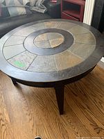 Round coffee table with stone tile inlay shown from a three-quarter angle, highlighting the tabletop pattern and the wood grain floor beneath.