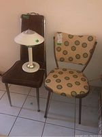 Two chairs and a table lamp arranged side by side on tile floor. One chair is dark brown vinyl with high back, the other is beige vinyl with circular green pattern. The lamp has a white shade with green details.