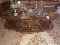 Full view of oval faux wood coffee table with glass top inserts, decorative carved legs, and storage cabinet doors with floral inlays, placed on patterned carpet.