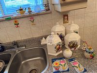 Set of four white hexagonal covered containers with colorful fruit designs including pears and grapes, displayed on kitchen counter near sink with additional decorative items such as glass candle holders and small ceramic pieces.
