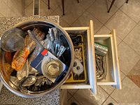 Two drawers filled with assorted cutlery and kitchen utensils next to a large metal bowl containing kitchen tools, strainer, scissors, and packing tape.