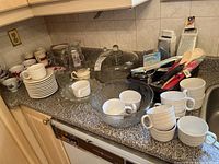Countertop with various white ceramic cups, plates, saucers, clear glass bowl, covered cake plate and pitcher.