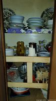 Shelves with stacks of various plates, bowls, pitchers, and mugs including large decorative china plates and amber glass pitcher