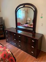 Front and side view of a dark wood dresser with six drawers and curved metal handles, topped with matching large mirror with rounded arch frame.