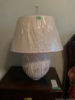 Ceramic lamp with white textured base featuring floral detail and cream fabric shade wrapped in plastic, placed on wooden surface.