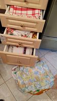 Four drawers partially open containing folded kitchen linens including dish towels, tablecloths, and placemats, plus linens on the floor