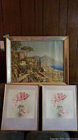 Three framed prints displayed against a wood-paneled wall. Large coastal landscape print at the back with two smaller floral prints with pink flowers in front.