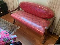 Front side angle of vintage wooden settee with tufted red vinyl seat and backrest covered by clear plastic.