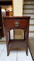 Front view of wood side table showing drawer with oval metal handle and open lower shelf.