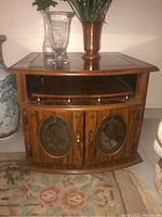Front view of faux wood side table showing top shelf with brass railing and two decorative glass inset doors with floral designs.