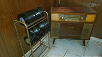 Photo of vintage Loewe Opta stereo console in wood cabinet, metal record stand with black vinyl records, placed in a corner with tiled floor and wood paneled walls.