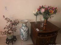 Photo showing large ceramic vase with floral and butterfly design beside faux cherry blossom tree on wooden stand. Also visible is a low wooden cabinet with clear and copper vases with artificial flowers.