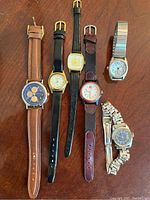 Six watches laid out on a wooden surface showing a variety of dial styles and bands including leather and metal.