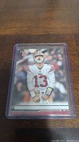 Photo of Brock Purdy rookie football card in a rigid plastic protective case on a wooden surface.