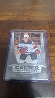 Photo of Jack Hughes Calder Front-Runners rookie hockey card in protective sleeve on wooden surface.