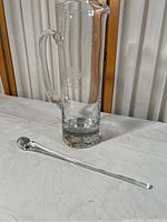 Full view of tall clear glass pitcher with handle and matching glass stirrer lying beside it on a white surface.