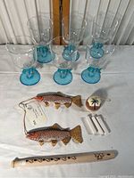 Photo showing six clear plastic cups with blue fish decorations, two wooden fish with detailed painting, a small round wind chime with palm tree design and a white wooden wind pipe.
