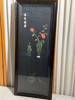 Full view of the tall black wood shadow box with soapstone carved figures inside, displaying a green vase with orange flowers and two other carvings on black background.