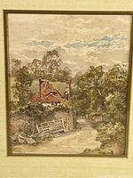 Close-up of framed vintage print showing country cottage with thatched roof, brick chimneys, surrounded by trees and a dirt roadside with white fence, in muted colors.