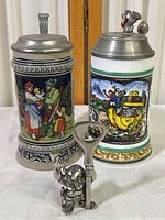 Two German beer steins side by side with metal lids. Left stein blue with family and cherub relief design. Right stein white glass with colorful horse and coach scene and a French Horn topper. Small metal novelty bottle opener figure in front.