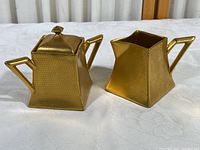 Front view of the gold sugar bowl with lid and cream pitcher showing their shape and honeycomb pattern on a white cloth background.