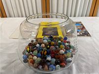 Clear glass bowl filled with assorted marbles with books partially visible behind.