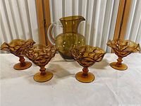 Set of five amber glass footed dishes and one amber glass pitcher with clear handle displayed on table.