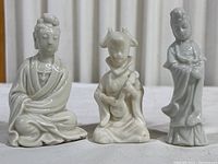 Group photo of three small white porcelain Far East style figurines on a white surface with a vertical blind background.