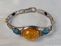 Front view of a silver bracelet showing a large oval yellow amber centerpiece flanked by two turquoise stones, set on an openwork silver band with smaller multi-colored amber stones.