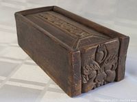 Side view of the antique carved wooden tea box showing the detailed carved motif on the end and slide top lid with central carved panel.