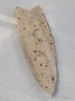 Front side view of flint spearhead showing serrated edges, pointed tip and notched base, light beige color with dark spots.