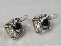 Front angled view of both square cushion-cut clear Swarovski crystal earrings in silver-tone metal prong setting on white textured fabric background.